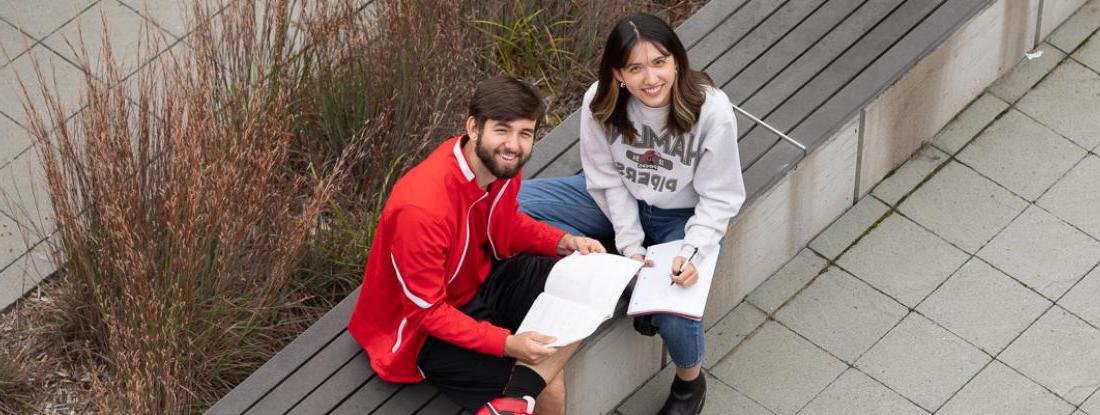 两个pg电子游戏的学生拿着书坐在校园外面，对着镜头微笑